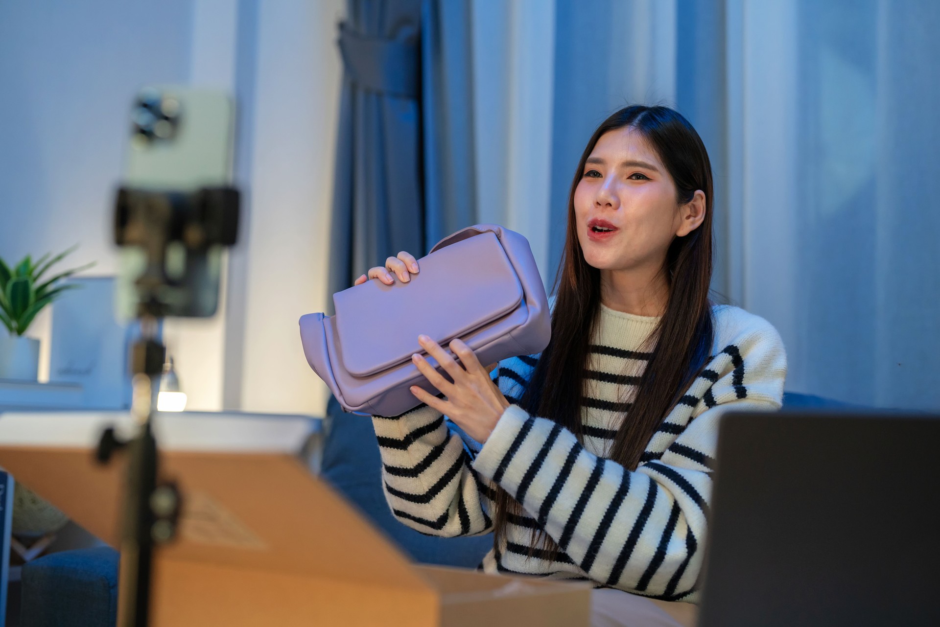 Asian female influencer livestreaming from her living room, selling stylish women's shoulder bags via an online platform using a smartphone. Showcasing modern e-commerce, digital marketing, and engaging with her audience in real time.